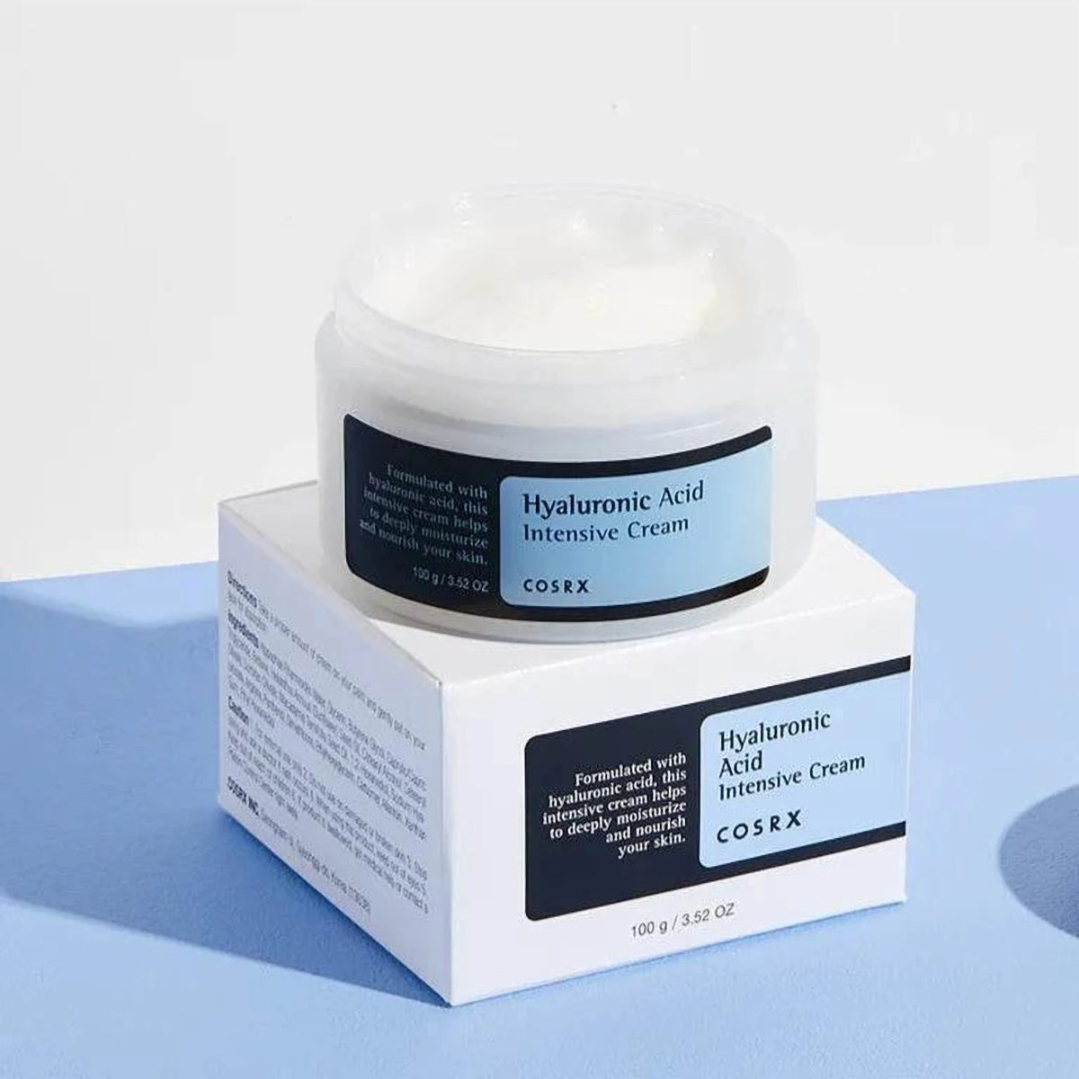 Pot ouvert de la crème COSRX Hyaluronic Acid Intensive posé sur sa boîte blanche, fond bleu