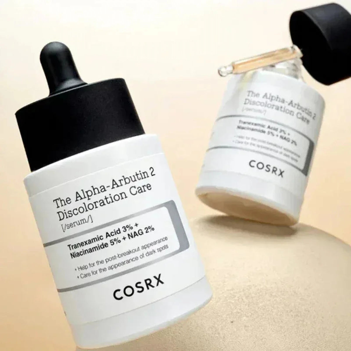 Deux flacons du sérum COSRX The Alpha-Arbutin 2 Discoloration Care sur fond beige, pipette sortie