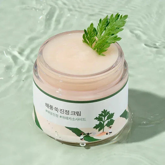 Pot ouvert de la crème Round Lab Mugwort montrant une texture rose pâle, feuille d'armoise posée dessus