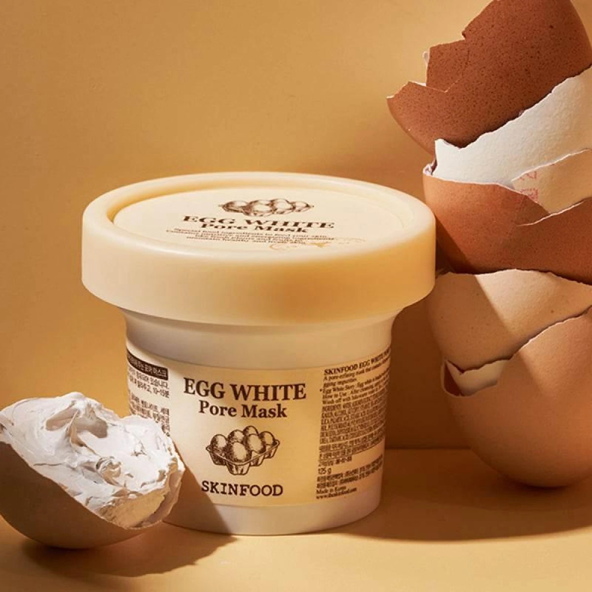 Pot du masque SKINFOOD Egg White Pore Mask entouré de coquilles d'œufs brunes sur fond ocre