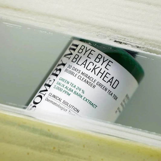 Flacon du nettoyant Some By Mi Bye Bye Blackhead posé derrière une vitre, texture verte en bas