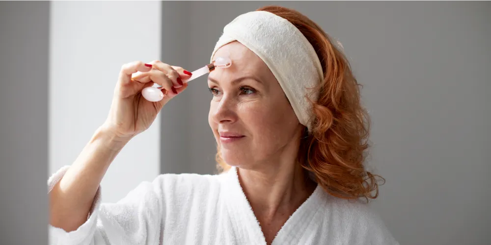 Femme utilisant un rouleau de jade sur son front pour un massage du visage, accessoire de skincare, avec un bandeau et un peignoir blanc.