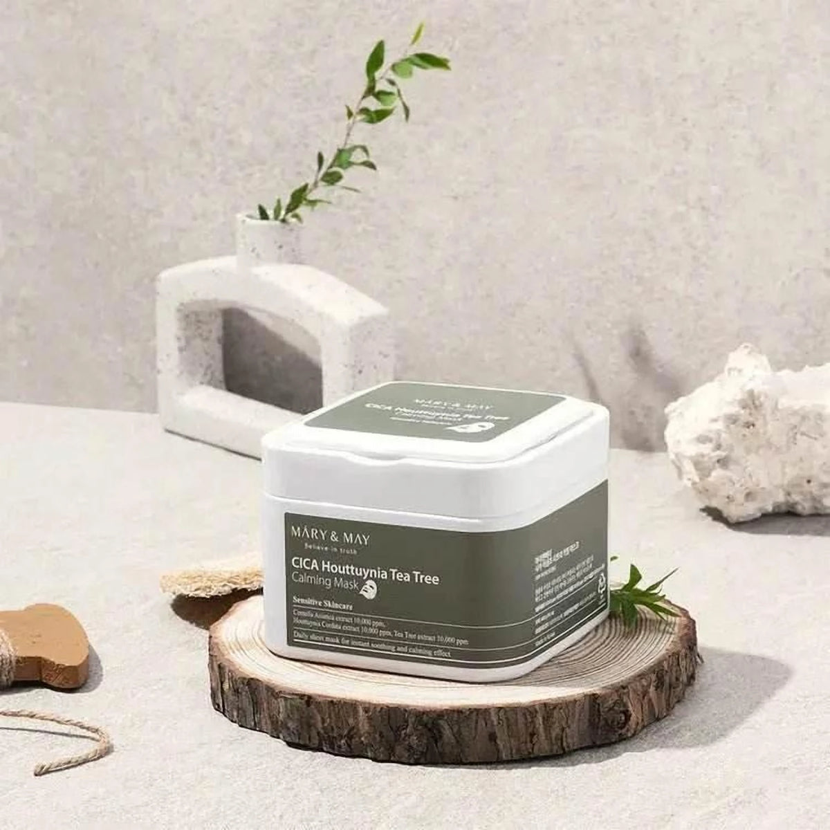 Boîte des masques Mary&May Cica Houttuynia Tea Tree posée sur un rondin de bois avec un brin vert
