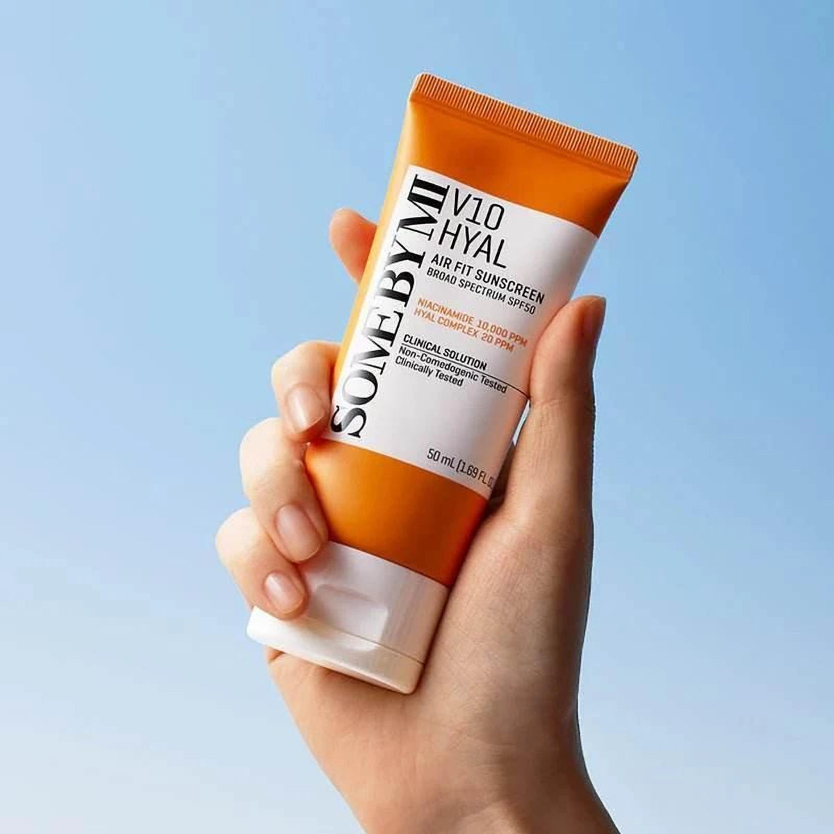 Main tenant le tube orange de la crème solaire Some By Mi V10 Hyal Air Fit devant le ciel