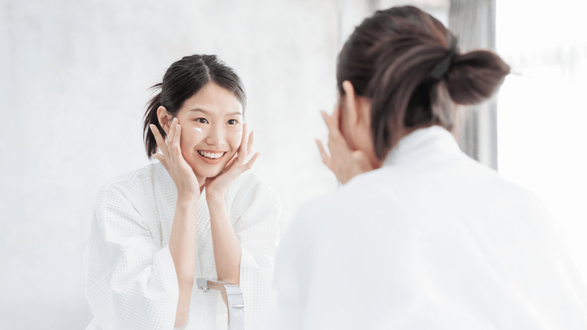 Femme souriante appliquant de la crème contour des yeux devant un miroir, portant un peignoir blanc