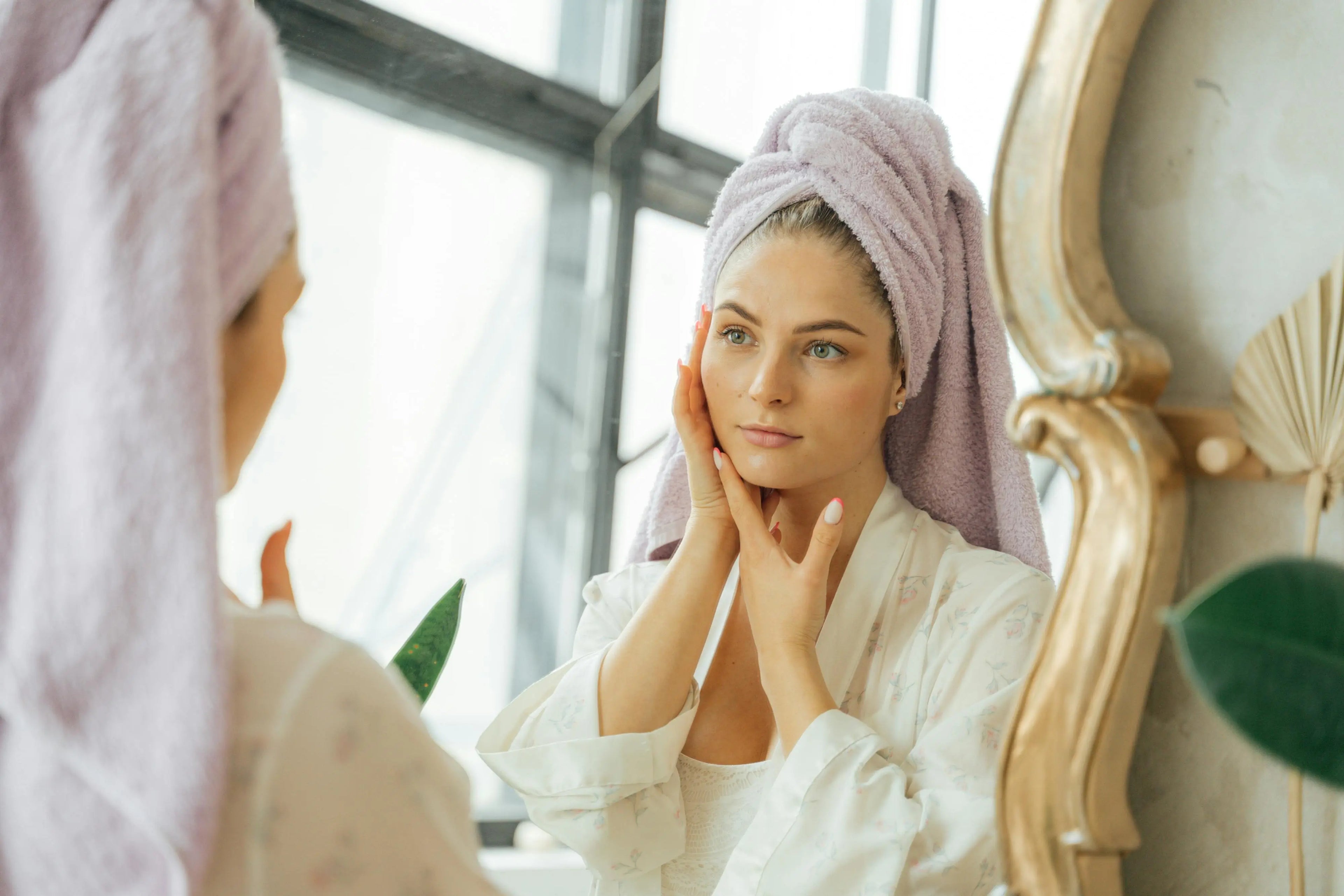 Femme avec une serviette sur la tête appliquant une crème ou lotion sur son visage devant un miroir, dans une routine de soin de la peau après la douche