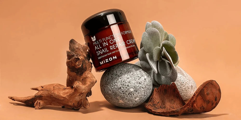 Pot de crème Mizon All In One Snail Repair Cream posé sur des galets et du bois flotté, soin coréen à la bave d'escargot pour la réparation cutanée.