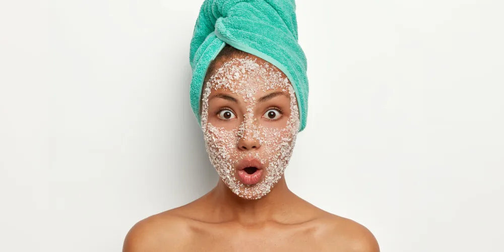 Femme avec un masque exfoliant granuleux sur le visage, serviette verte enroulée sur les cheveux, exprimant la surprise, routine skincare et soin des pores.